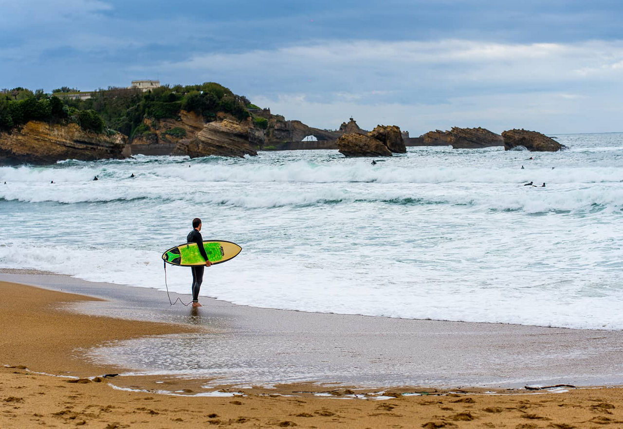 A surfer stands on the beach and looks out to sea, his surfboard in his hand