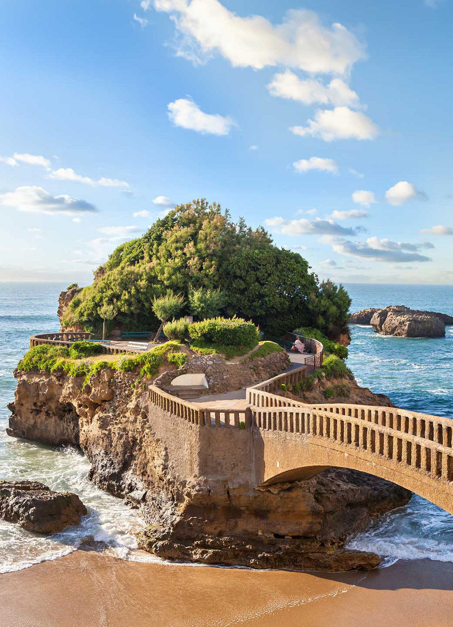 A droite de l'image, un pont de pierre qui mène à une petite île verte dans la mer.