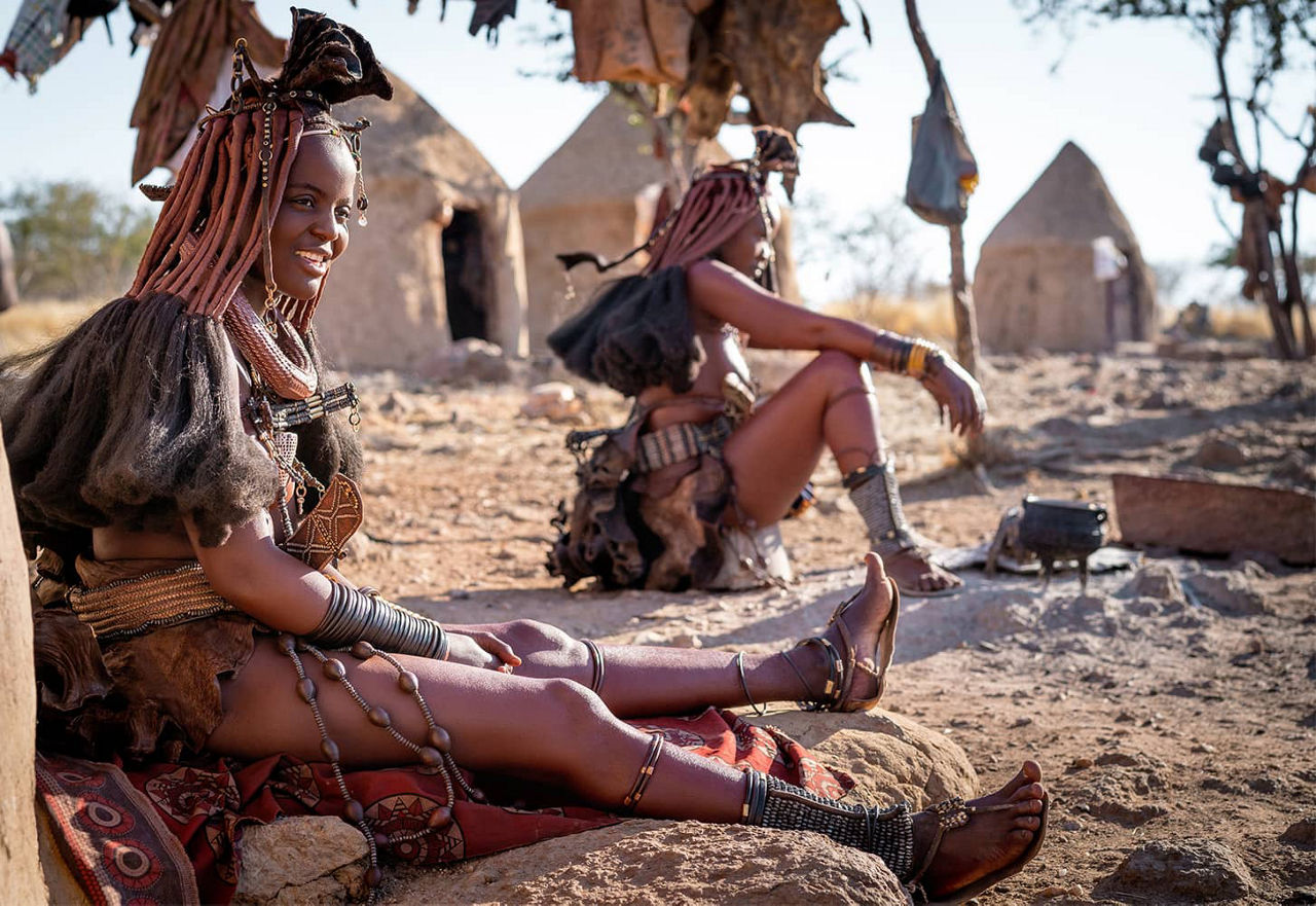 Zwei am Boden sitzende Himba-Frauen vor traditionellen Häusern